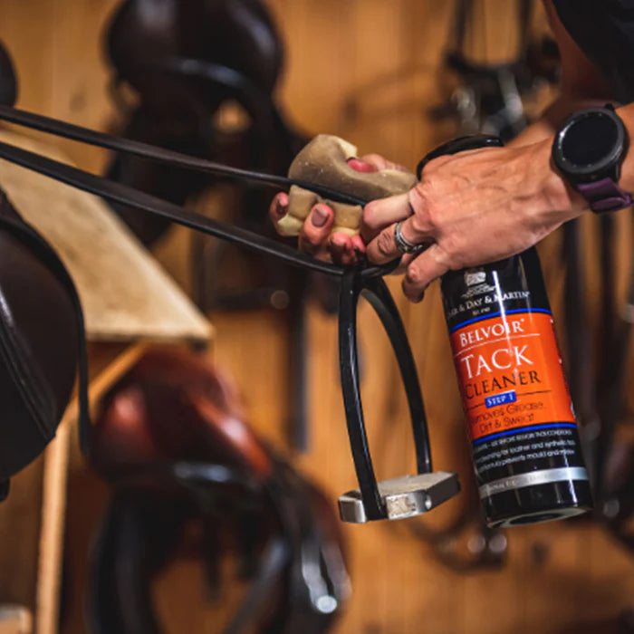 Person cleaning stirrup leathers with Belvoir Tack Cleaner in a stable setting.