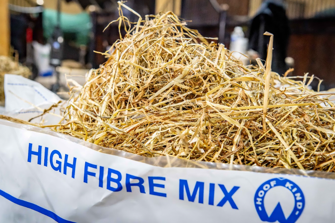 A white bag labeled 'HIGH FIBRE MIX' with blue text and a circular logo, filled with golden-yellow dried haylage in the foreground, photographed in a real indoor setting with a blurred background.