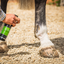 Person applying a green bottle of hoof care product to a horse's hoof on a gravel surface.
