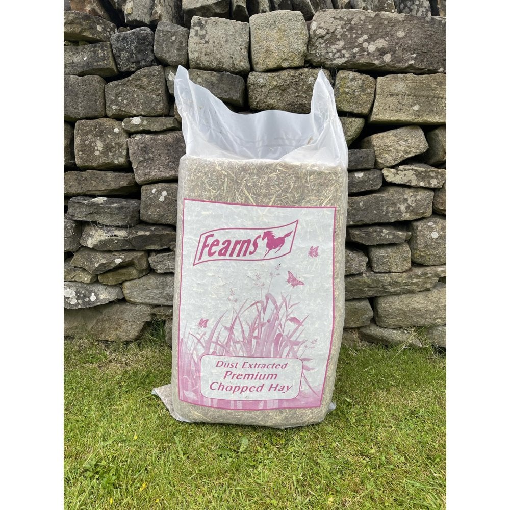 A large bag of Fearns Dust Extracted Premium Chopped Hay standing on grass in front of a rustic stone wall.