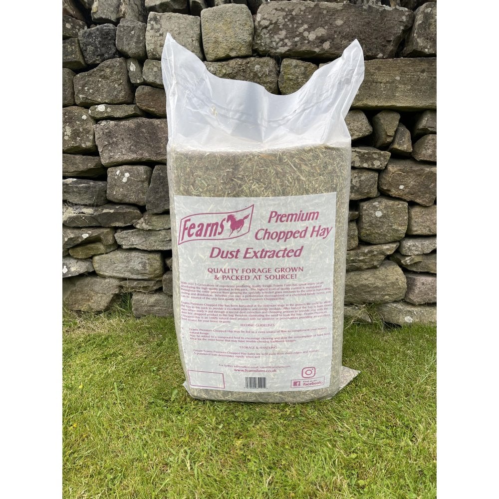 A large white plastic bag of Fearns Premium Chopped Hay, dust extracted and granulated, standing on green grass in front of a rustic stone wall.